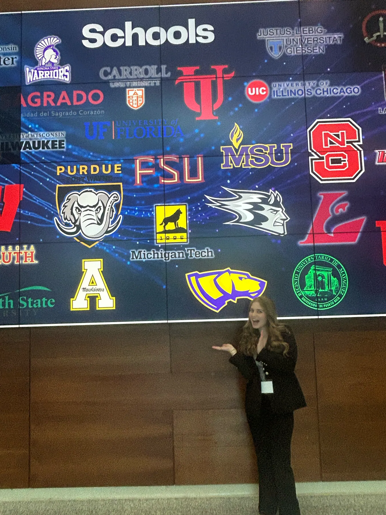 An attendee gesturing at a video wall of university logos titled Schools.